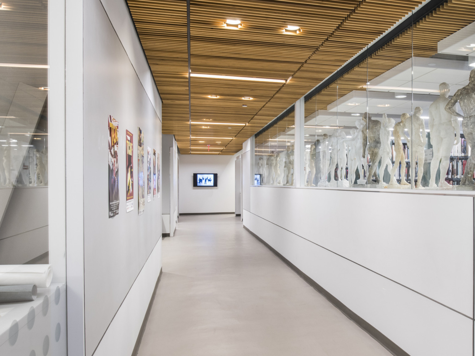 Hallway with wood ceiling and grey walls. Paper hangs on left wall, the right wall has a glass top displaying sculptures
