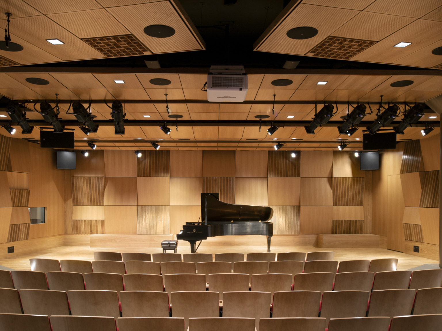 Auditorium with wood stage and seating, matching acoustical panels on stage walls and above, black light fixtures hang down