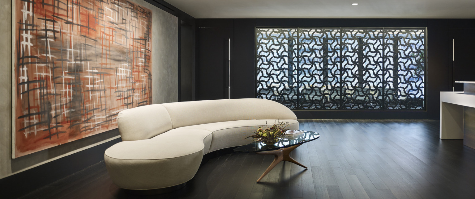 Curved white couch with glass coffee table in wood room with abstract art behind. Back wall has organically patterned panels