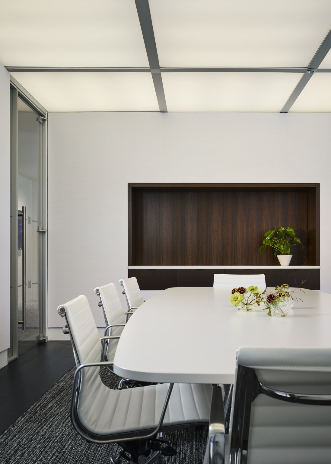 White conference room with long table and chairs. Recessed wood wall section, opposite, with white counter. Light panel above
