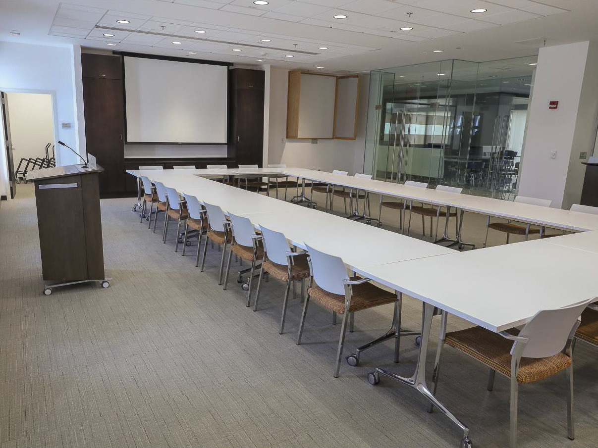 Conference room with grey carpet and long white tables and chairs arranged in a rectangle. Glass entry doors back right.
