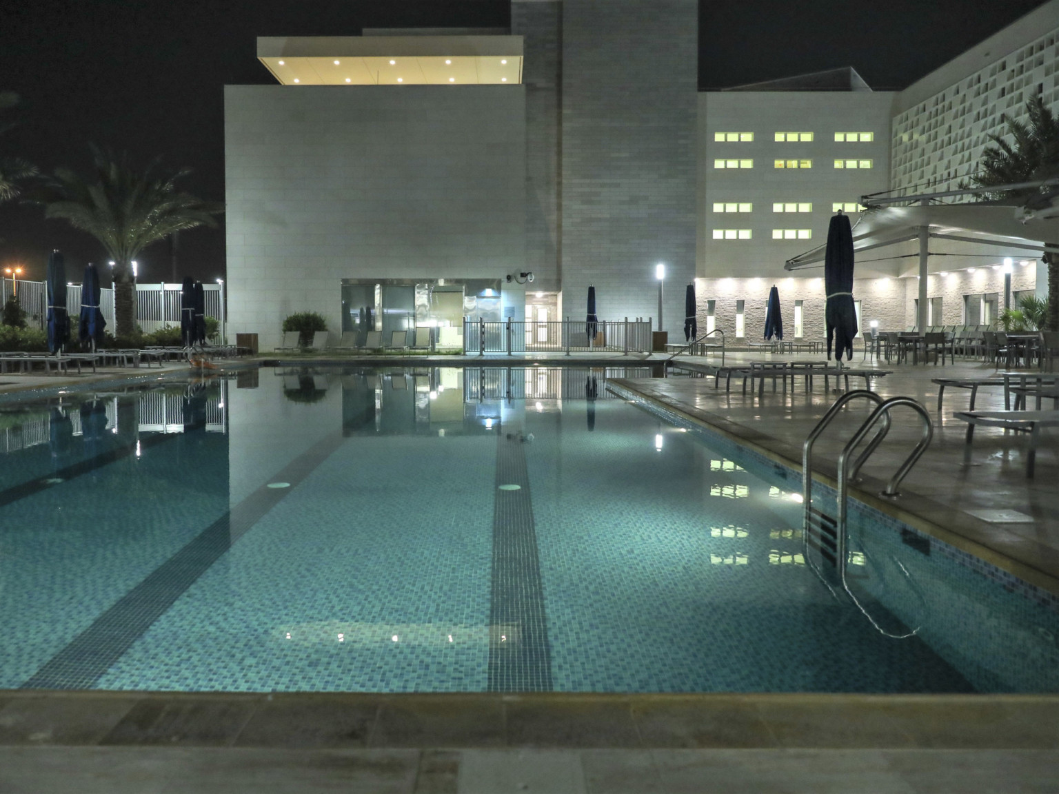 Outdoor swimming pool at night with lounge chairs and umbrellas right. Behind is a white building with covered roof terrace
