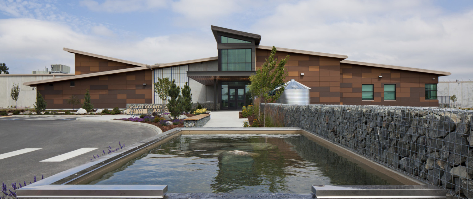 A stone building with angled glass central entryway. A path from the doors leads to a water feature next to a stone gabion