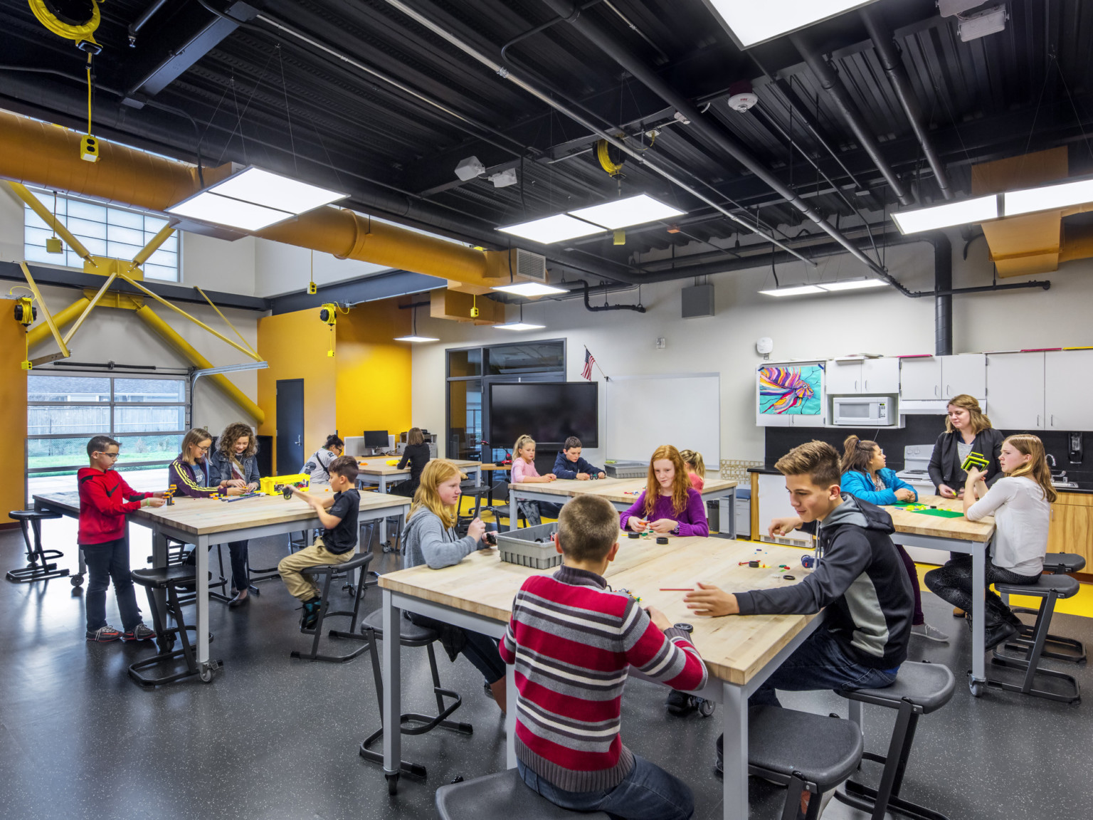 Wood topped tables with flexible seating in white classroom with yellow accents. The black ceiling has exposed pipes and duct