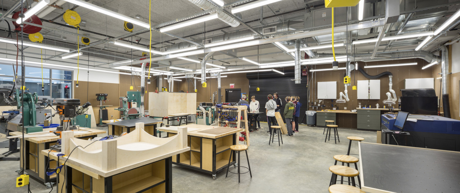 Large woodshop with equipment and projects. Exposed ductwork and extensions cords hang above