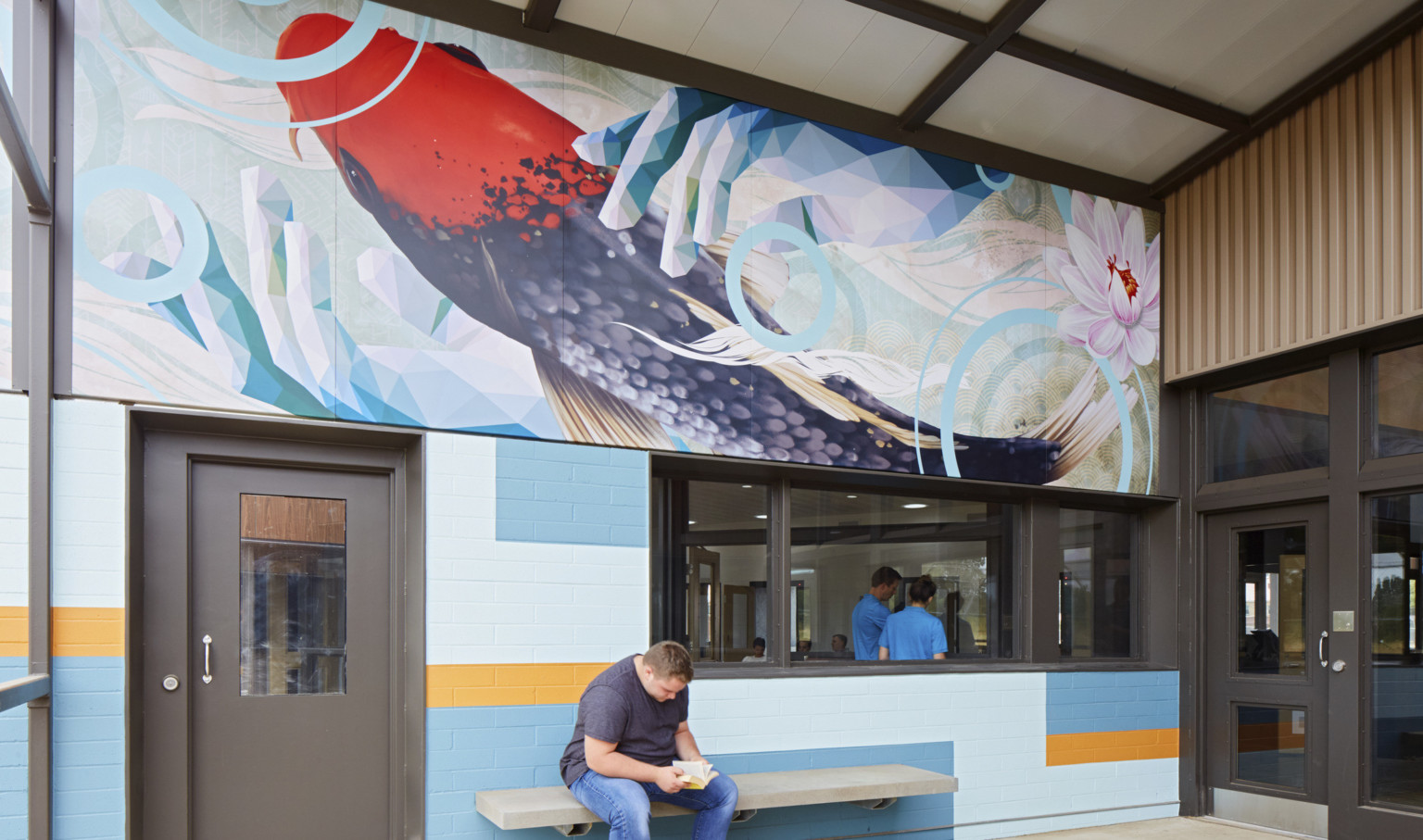 A grey bench against a blue and yellow colorblock wall with a window into an office. Above is a mural of a koi fish