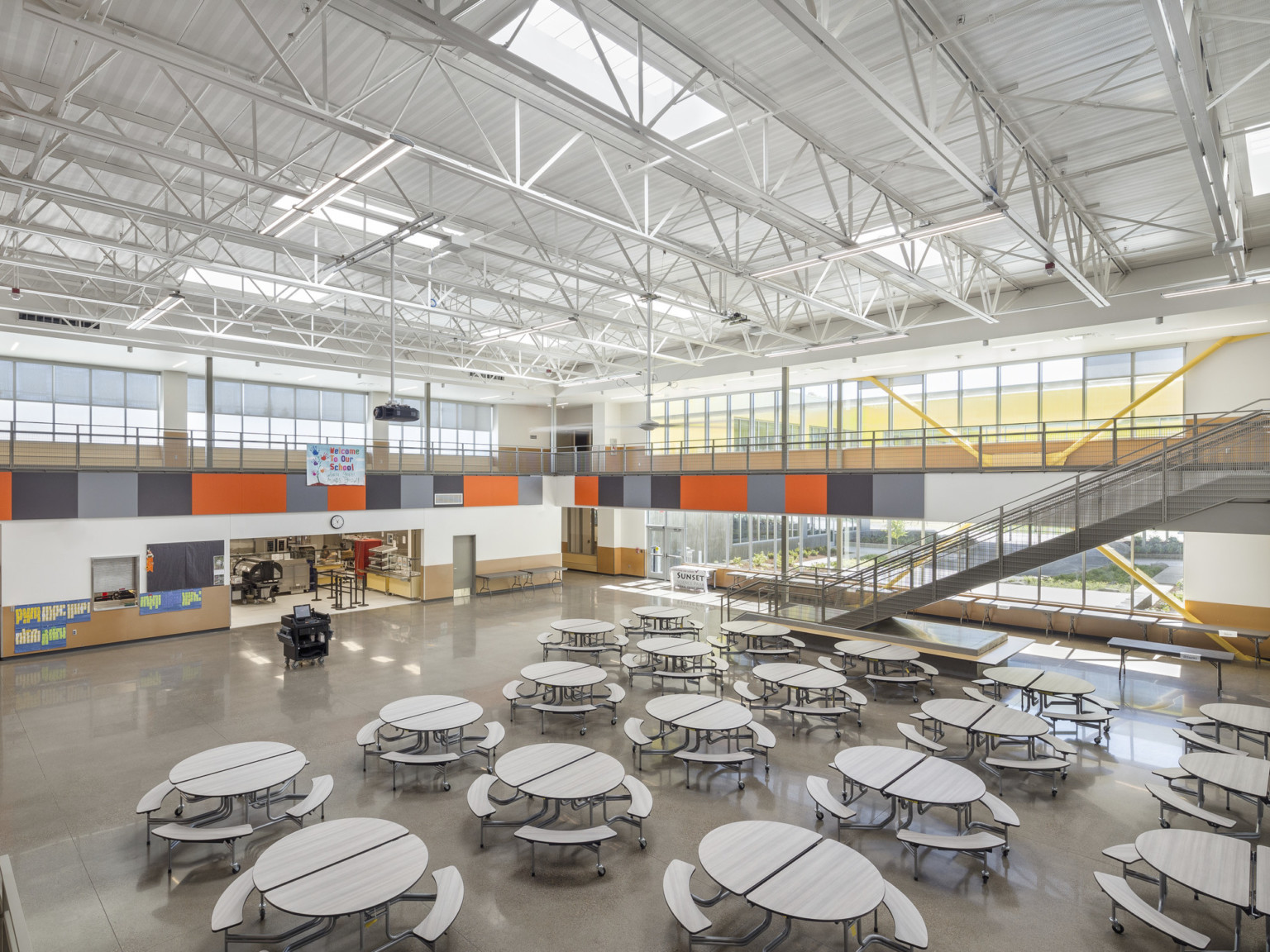Double height white cafeteria with orange accents. Windows along far wall. Round folding tables on ground level. Kitchen left