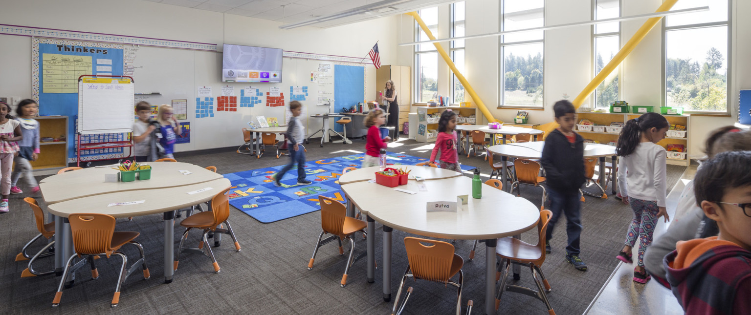 Classroom with yellow exposed beams at angles by windows, right. Oval kid sized tables and chairs around blue alphabet carpetClassroom with yellow exposed beams at angles by windows, right. Oval kid sized tables and chairs around blue alphabet carpet