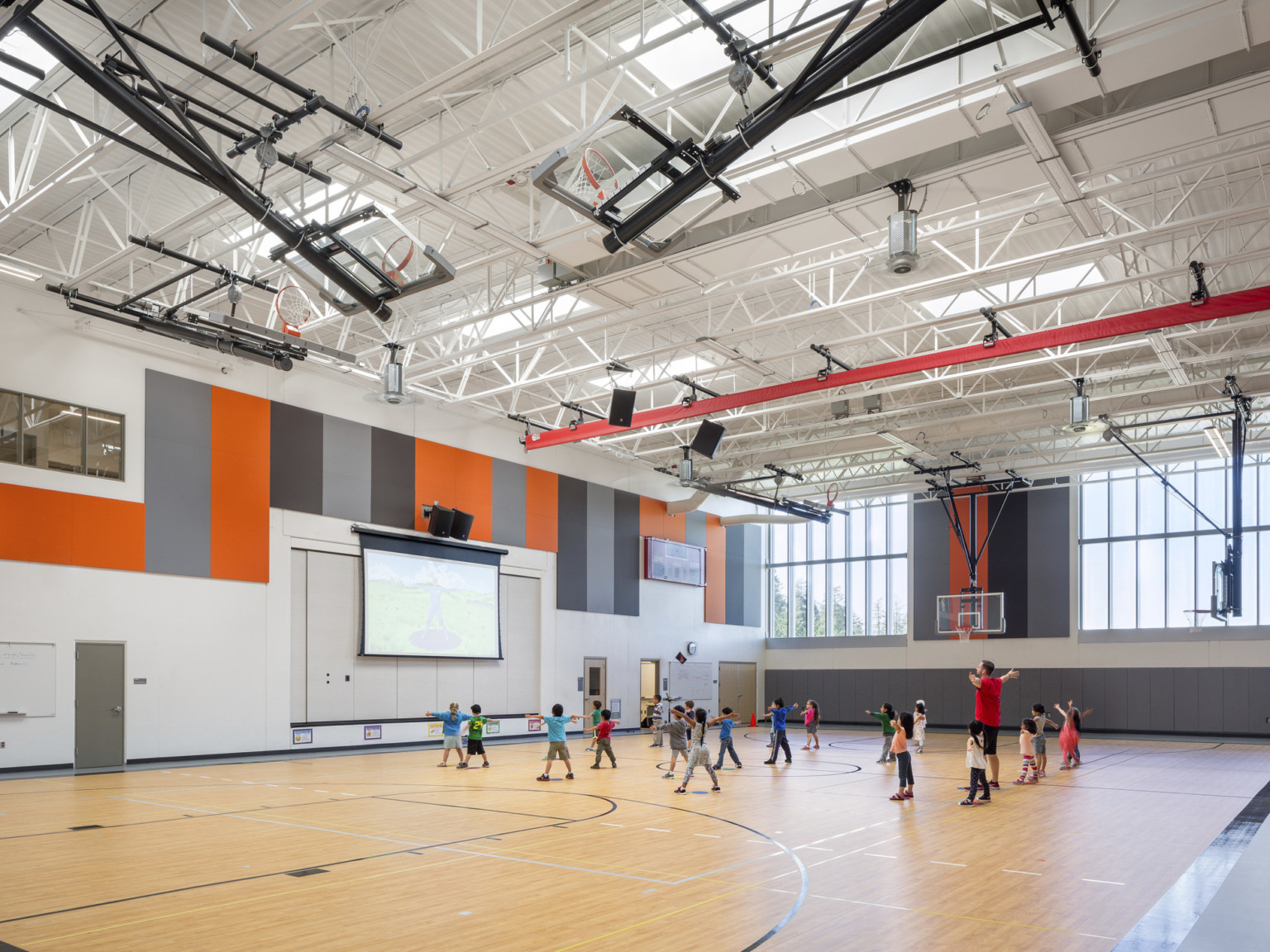 Double height gymnasium with exposed beams above. Wood basketball court with hoops raised. Projector screen at center left