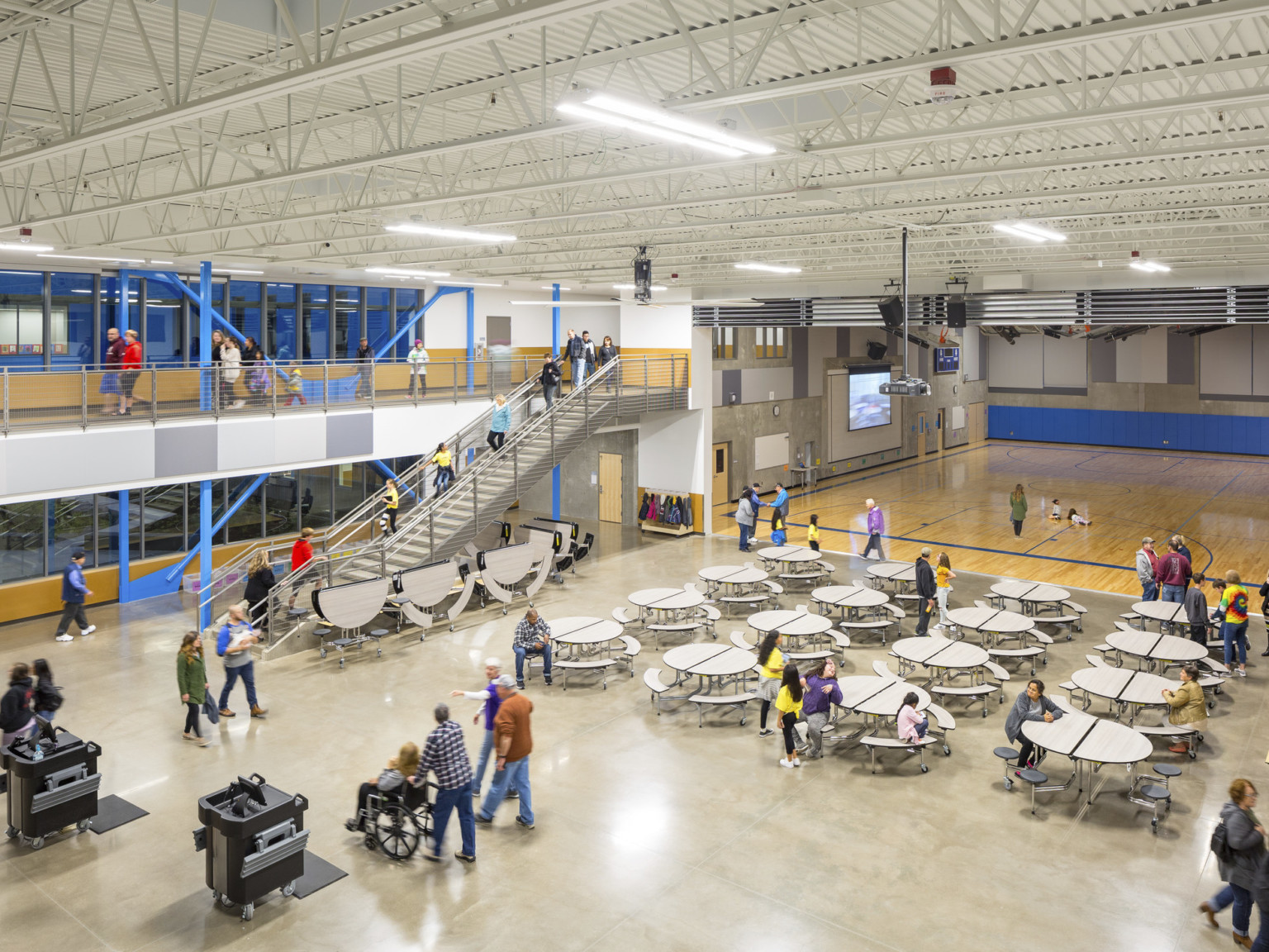 Cafeteria viewed from 2nd floor walkway facing gym. Rows of round folding tables with attached seating. Stairs on left wall