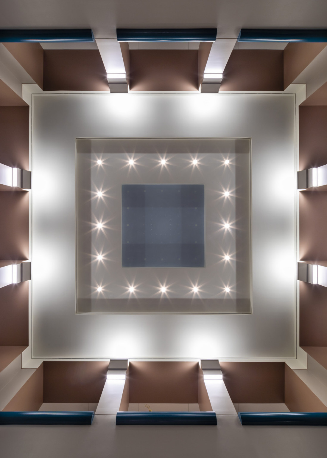 View of an interior ceiling from below. White columns meet at brown top edge of recessed square ceiling details and lights