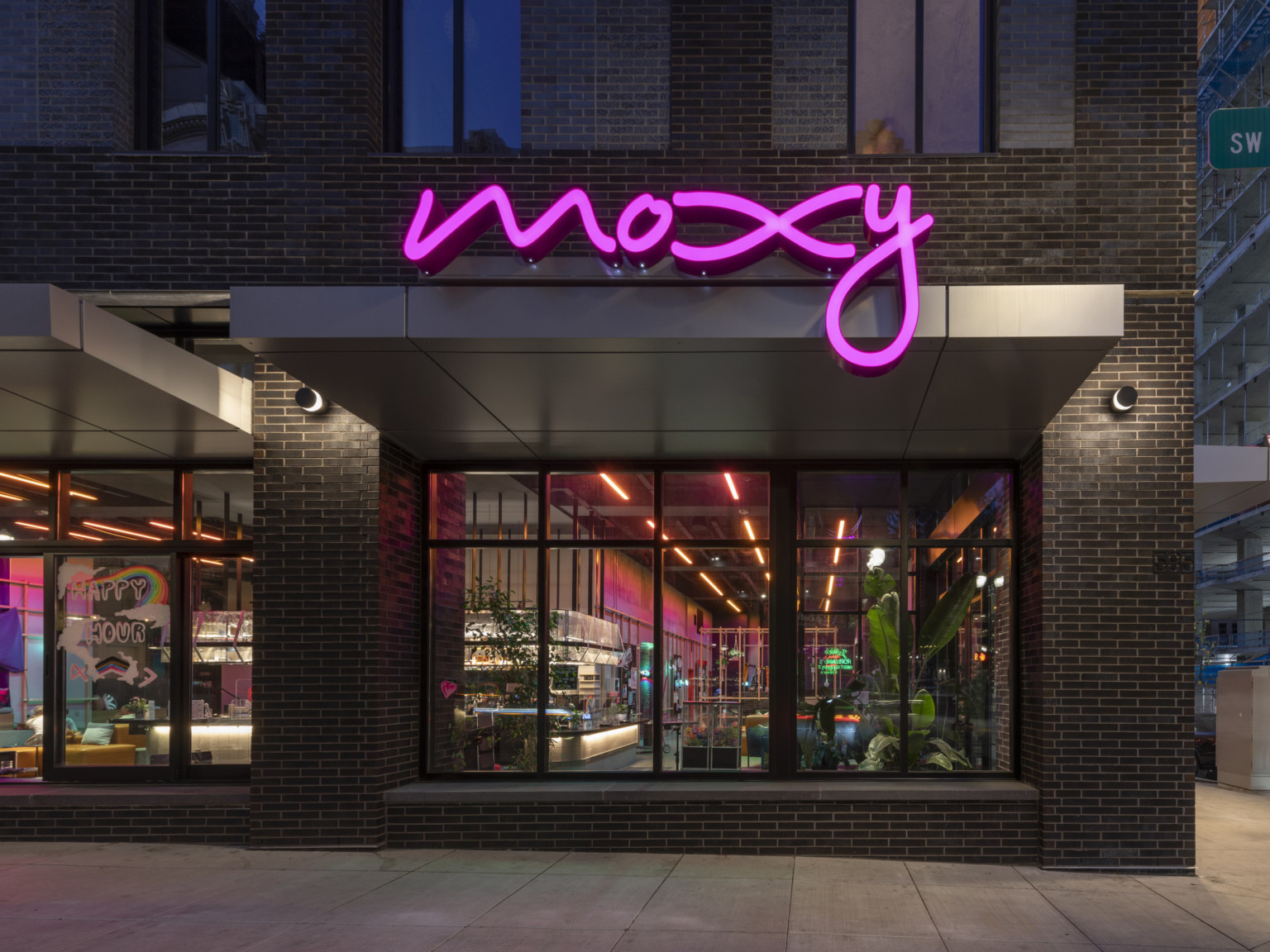 Exterior view from ground level corner at night, coffee shop can be seen inside through windows. Moxy pink sign illuminated