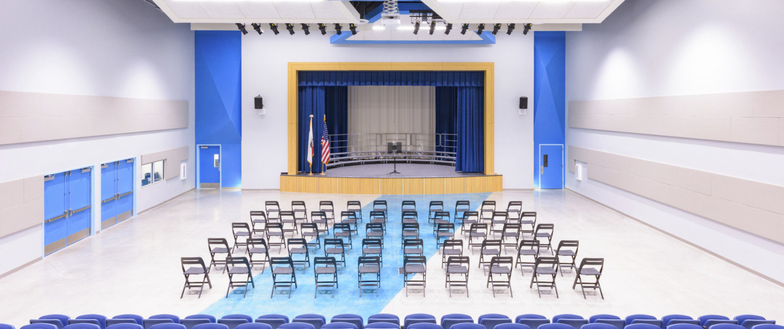 Auditorium with blue accents and doors. Folding chairs in front of blue chairs face stage with wood proscenium, blue curtains