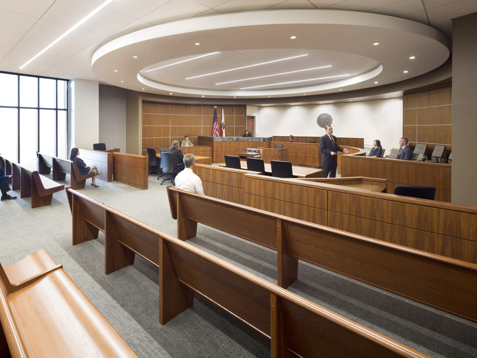 Courtroom with rounded judge's bench mirroring circular drop ceiling detail with recessed lighting. Wood accent wall on sides