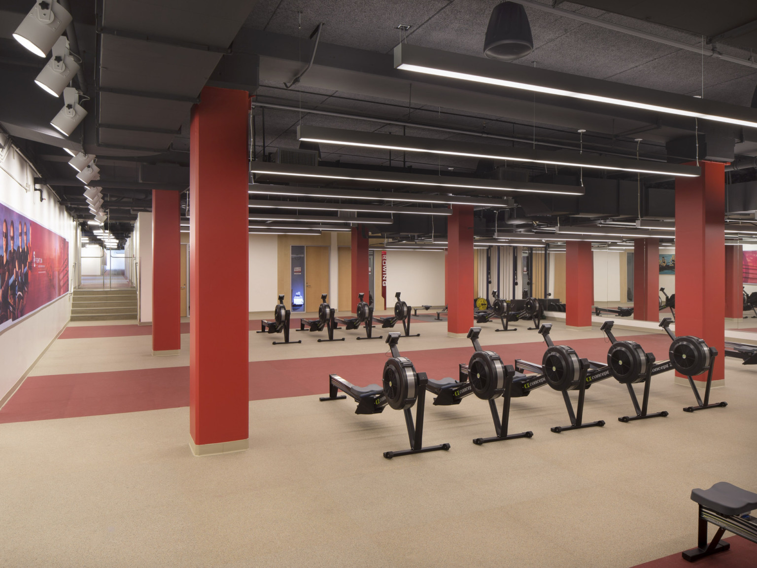 Training room with lines of rowing machines. White walls in room with orange columns to black ceiling with exposed ductwork