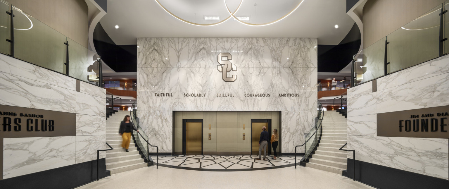 Marble side wall reading Founders Club frame curved double staircases and elevators in white room with round hanging lights