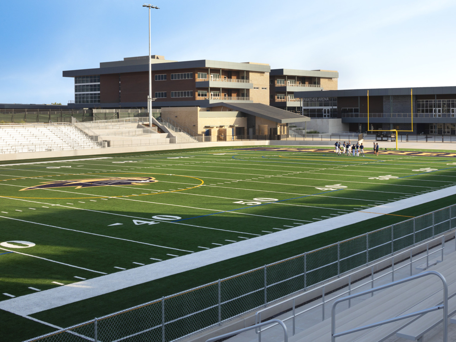 View across football field from sidelines. 2 story building at end zone behind main buildings to left. Metal bleacher benches
