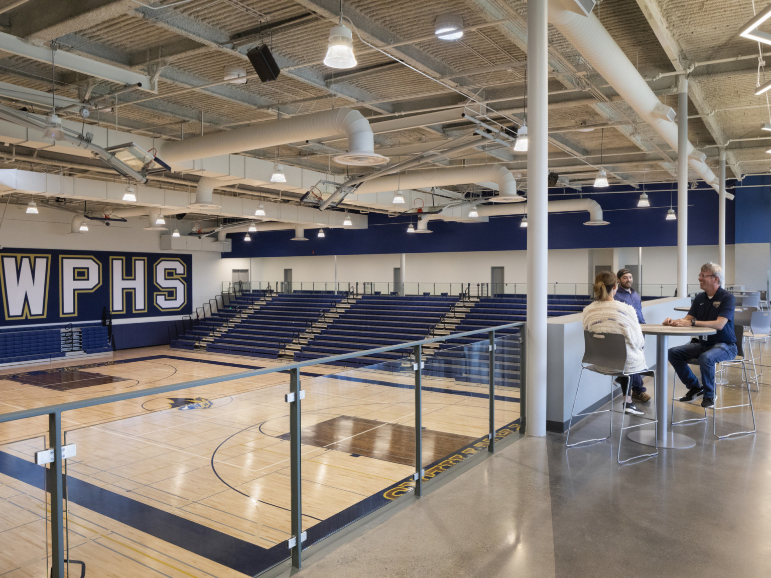 High top tables and chairs on 2nd floor balcony over double height basketball court with wood flooring and blue bleachers
