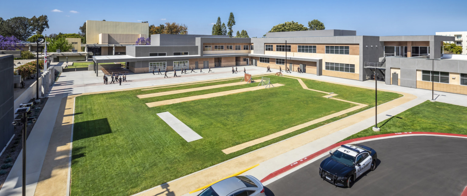 Corner aerial view of track and field with L shaped two story building in background, between which people train