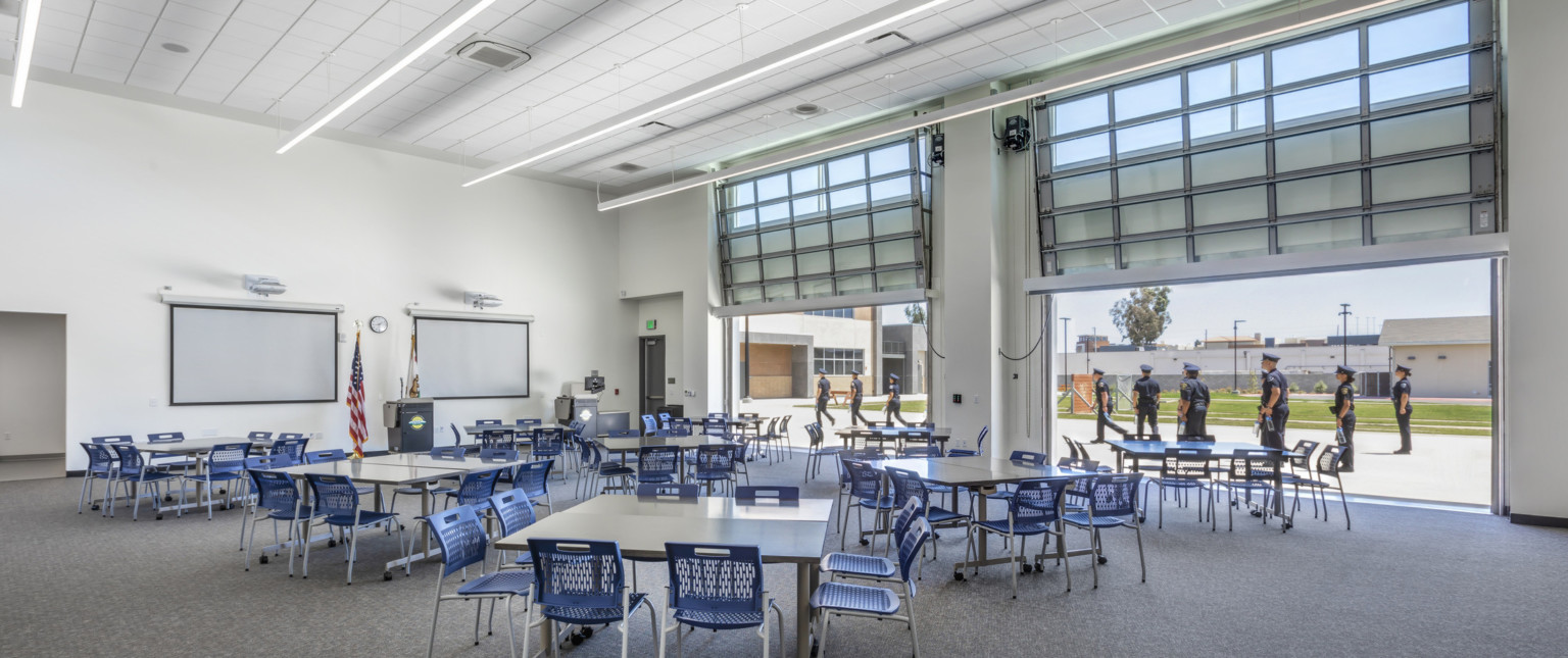 A large flexible work space with movable tables and chairs in front of a podium. Garage style doors are open to a field