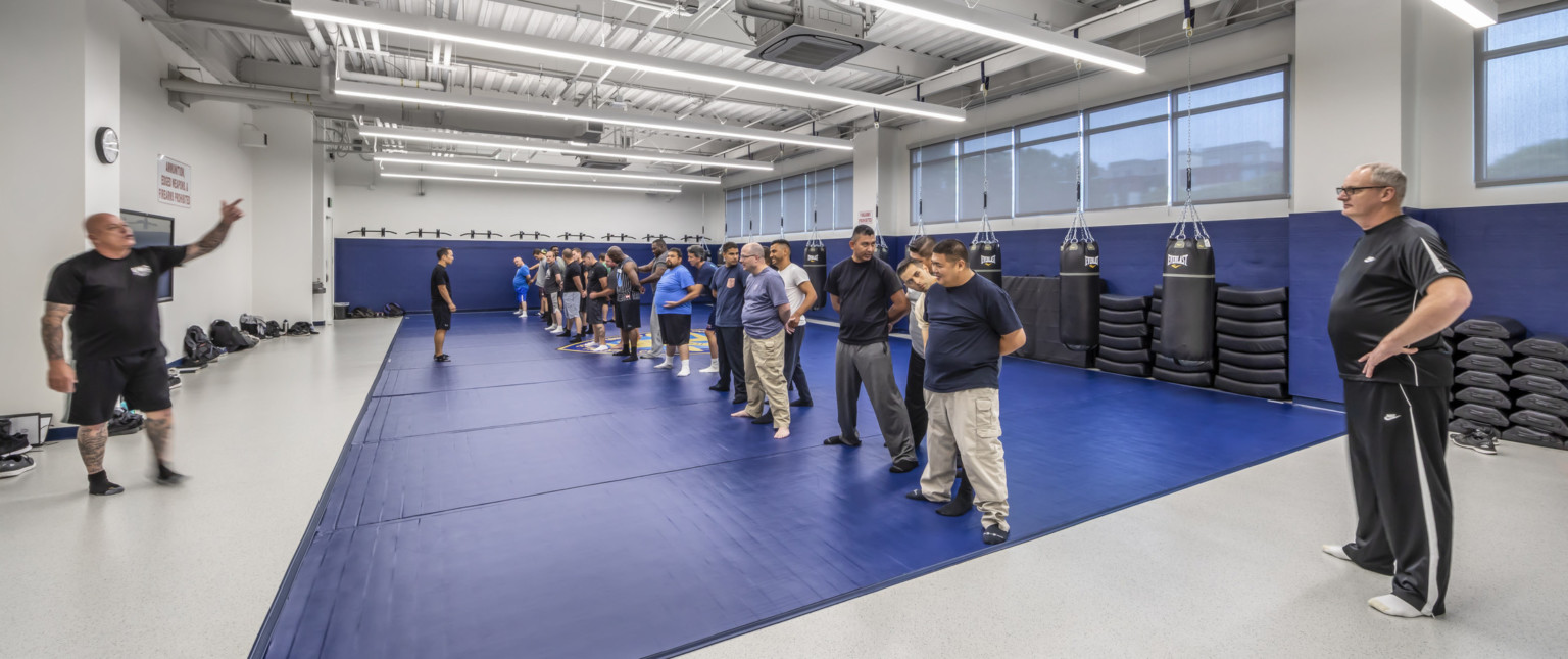 Exercise room with blue padding along floors and walls, men stand practicing at the center of the room with equipment behind