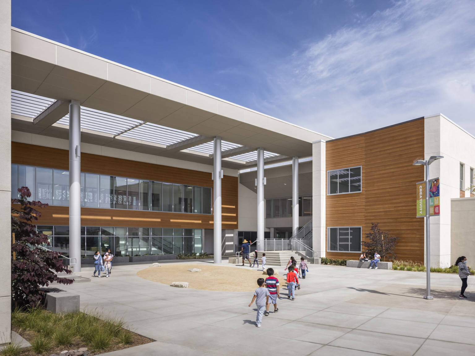Entry seen across plaza. White canopy with skylights covers entry. White walls with large windows and wood accents by steps