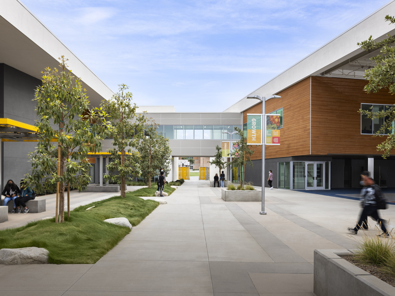 Exterior tree lined walkway between buildings. Grey walls with white at the top, and a wood panel accent on right upper wall