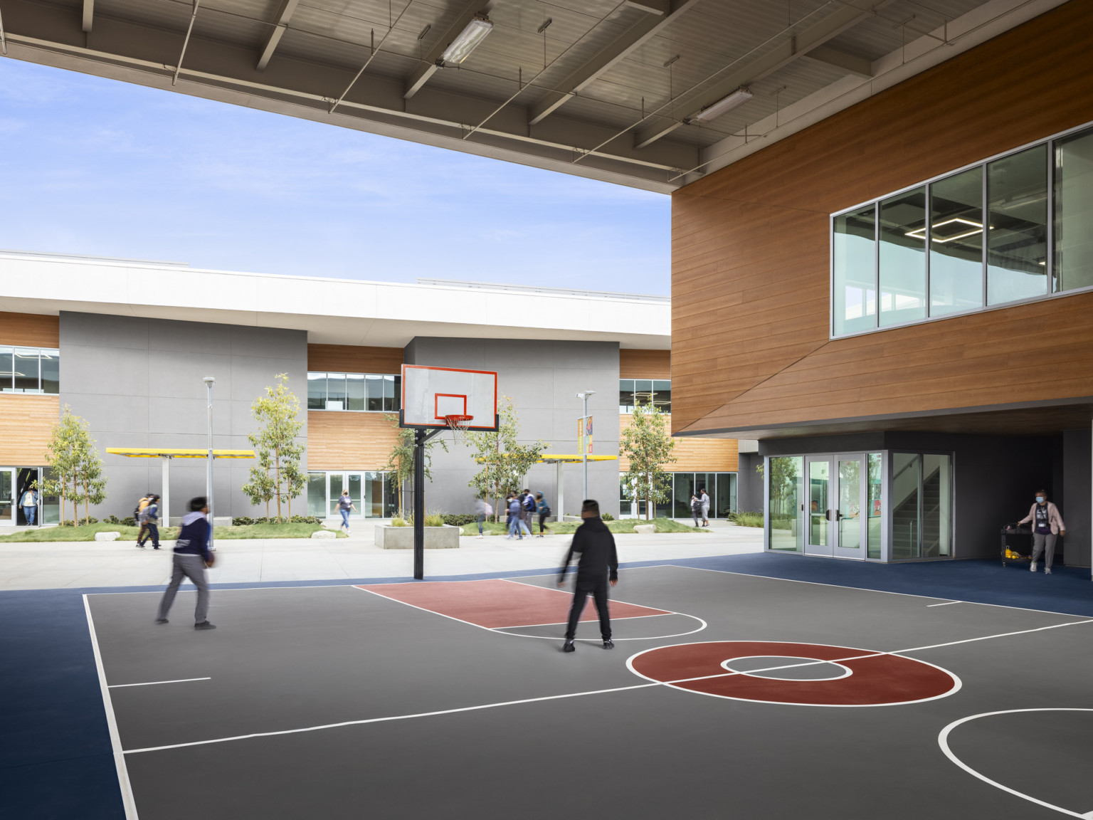 Exterior view of covered basketball court recessed between buildings. Right, 2nd floor, a wood panel wall with large windows