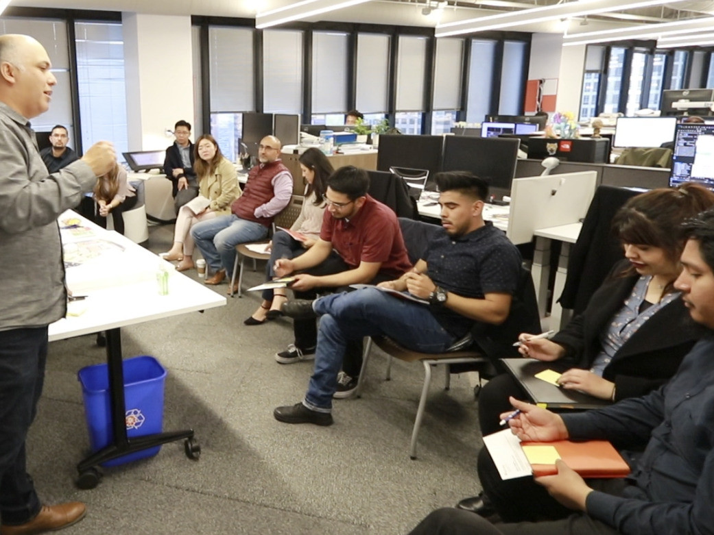 College students involved in design workshop sit in a row taking notes facing a man speaking College students involved in design workshop sit in a row taking notes facing a man speaking