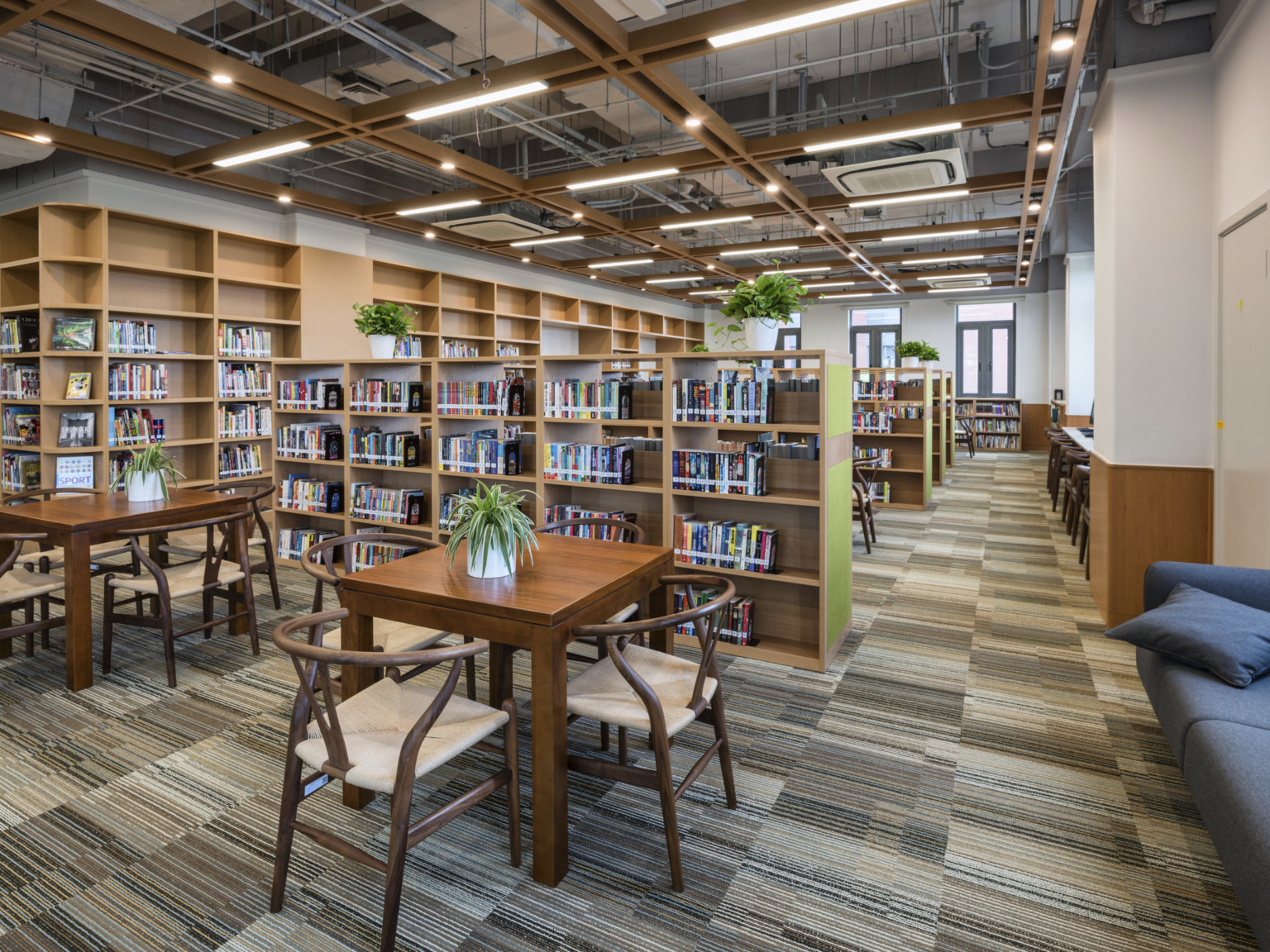 Carpeted library with flexible seating. Exposed pipes behind wood beams with recessed lighting. Wood bookshelves along wall