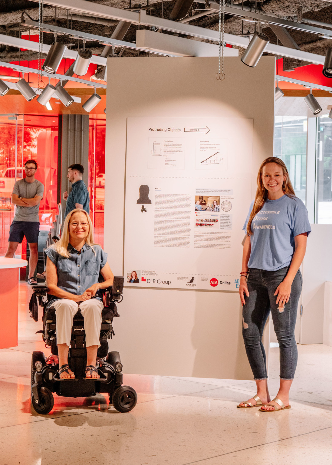 2 women in front of hanging white panel with information and charts in red room. Woman, left, sits in wheelchair.