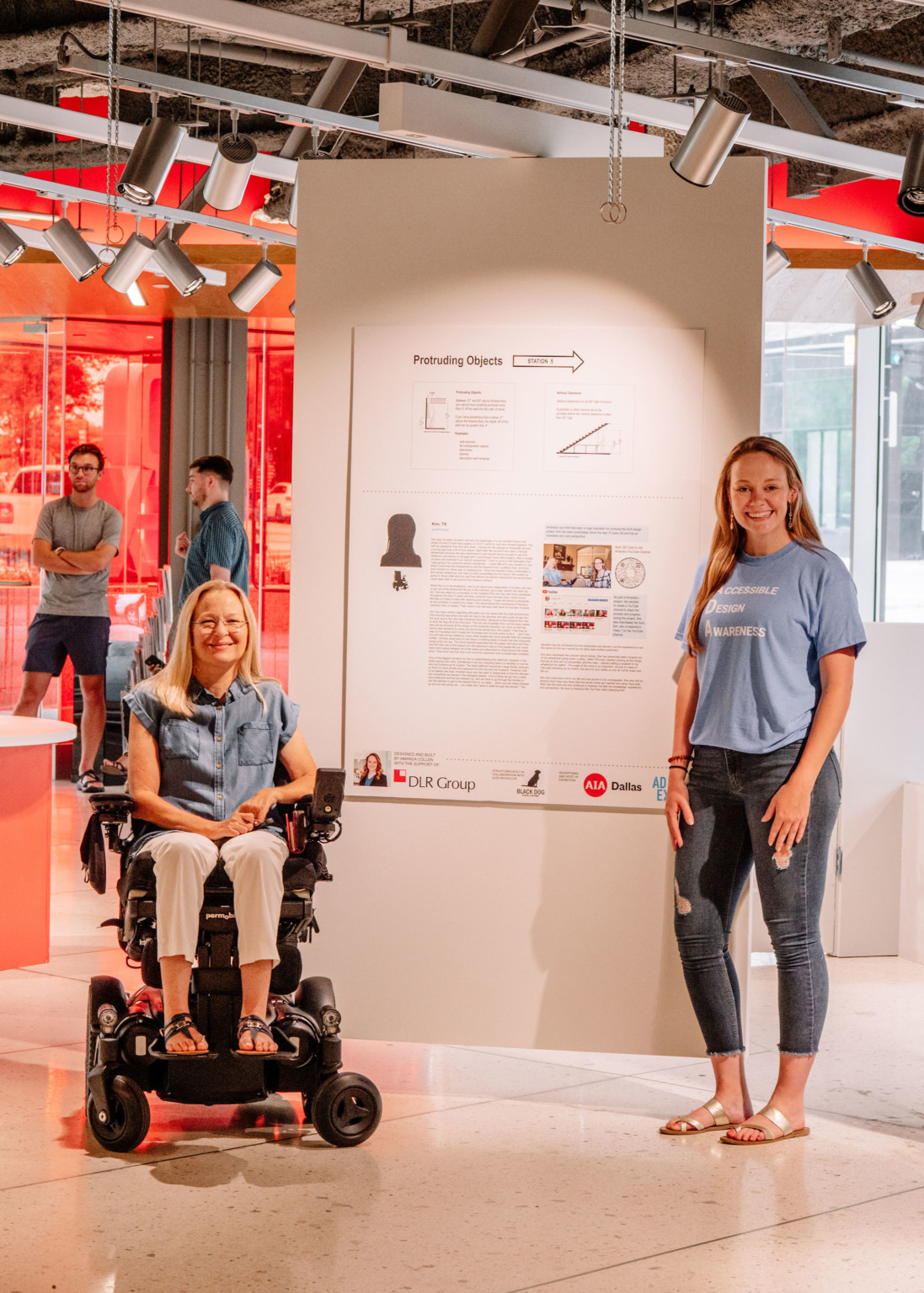 2 women in front of hanging white panel with information and charts in red room. Woman, left, sits in wheelchair. 2 women in front of hanging white panel with information and charts in red room. Woman, left, sits in wheelchair.
