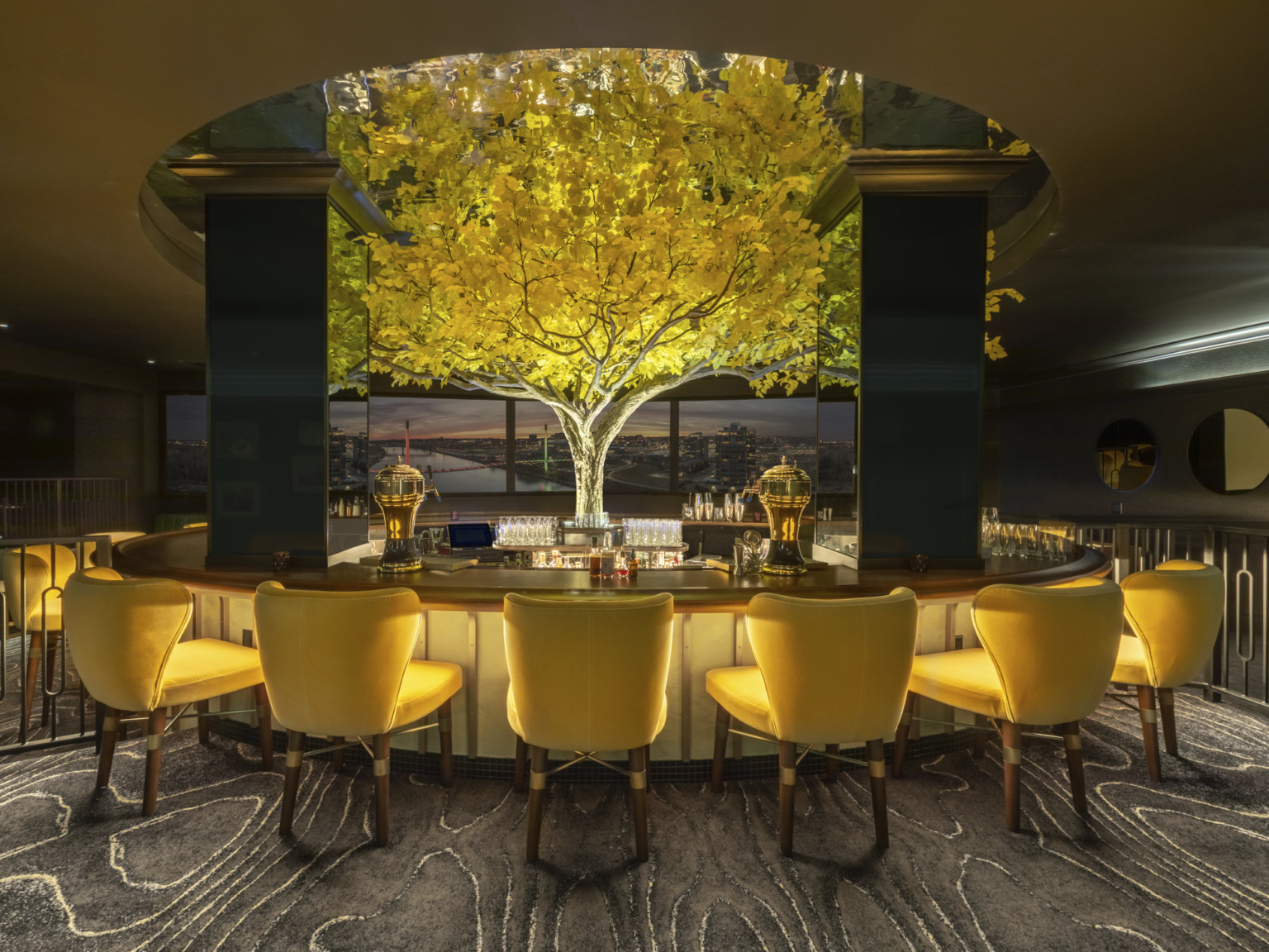 an illuminated yellow tree in the midst of in the round high top bar with yellow seats. Tree rises above round ceiling cutout