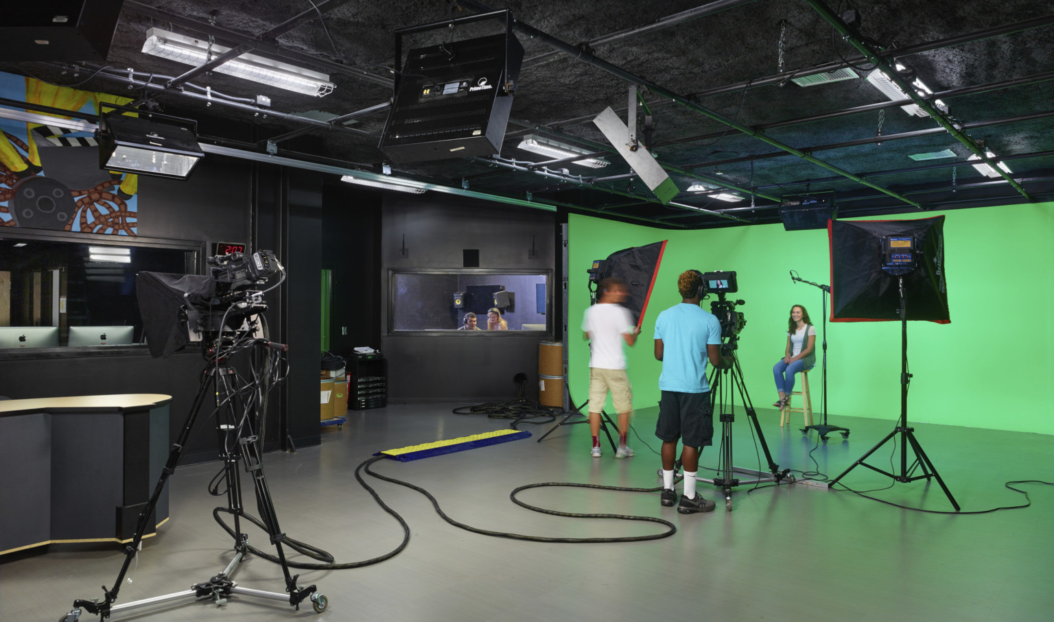 Students operate camera and lighting equipment in front of green screen in black room. Window, left, to editing room
