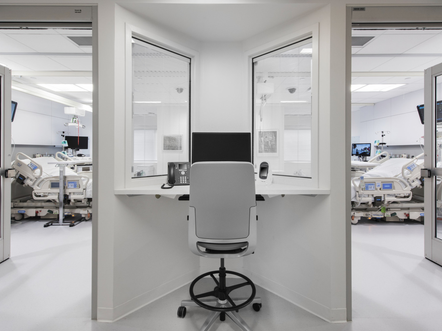 Desk with view into 2 hospital rooms, each with beds and screens. The white desk fits into a triangular space between doors