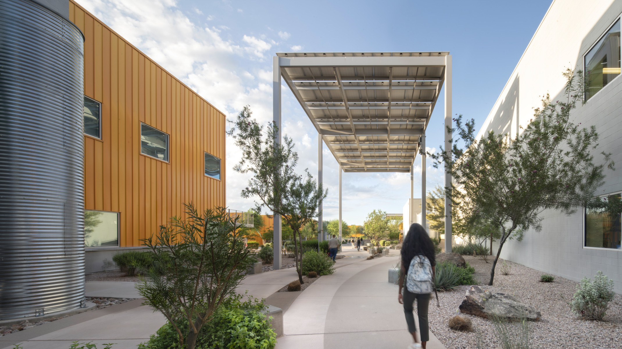 Student walking down an outdoor path with solar canopy above and an orange metal cladding building on left Student walking down an outdoor path with solar canopy above and an orange metal cladding building on left