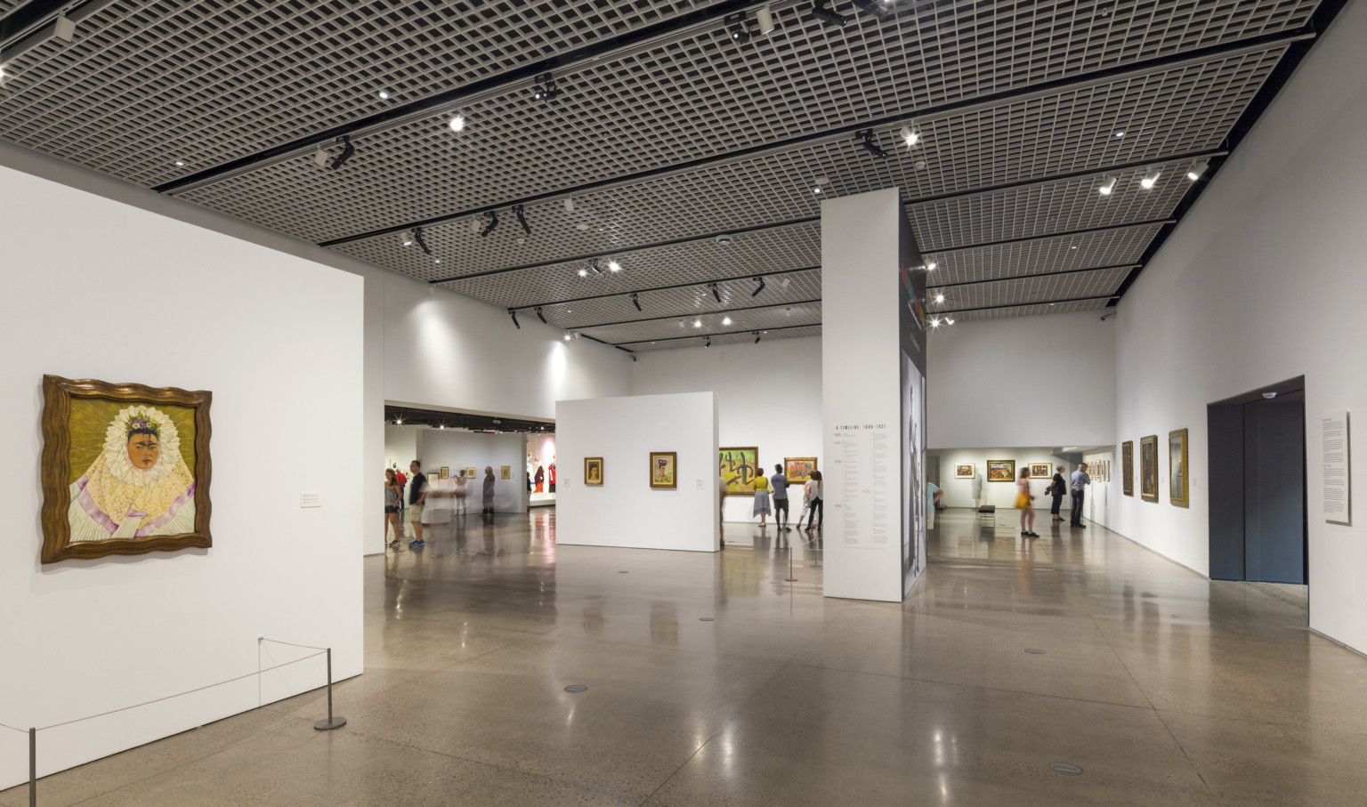 A steel grid with light fixtures hanging off spans the ceiling above a white gallery with paintings on the walls