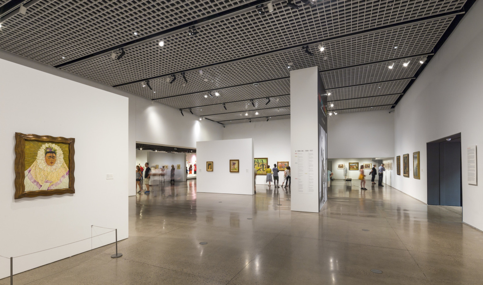 A steel grid with light fixtures hanging off spans the ceiling above a white gallery with paintings on the walls A steel grid with light fixtures hanging off spans the ceiling above a white gallery with paintings on the walls
