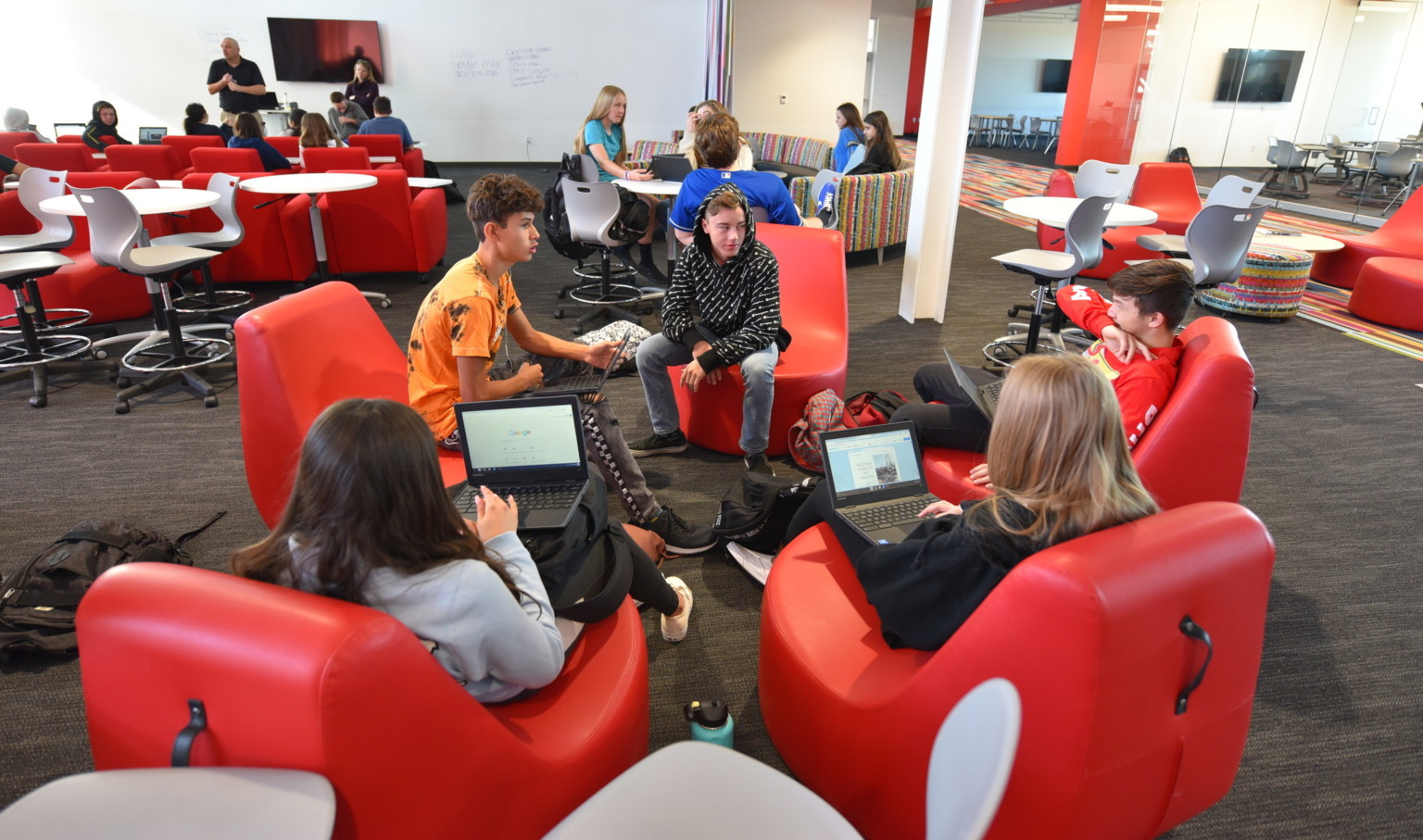 Students working on laptops in red ergonomic chairs in a large open learning space with flexible seating