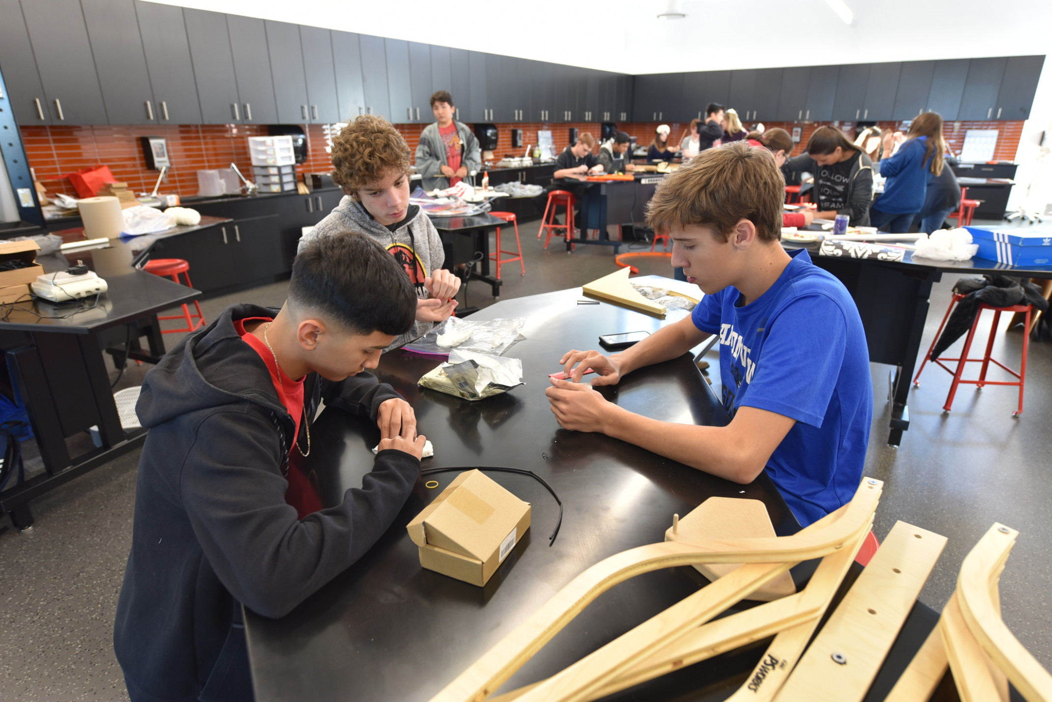 students working with wood pieces at a black rounded table in a classroom with black cabinets and orange tiled backsplash