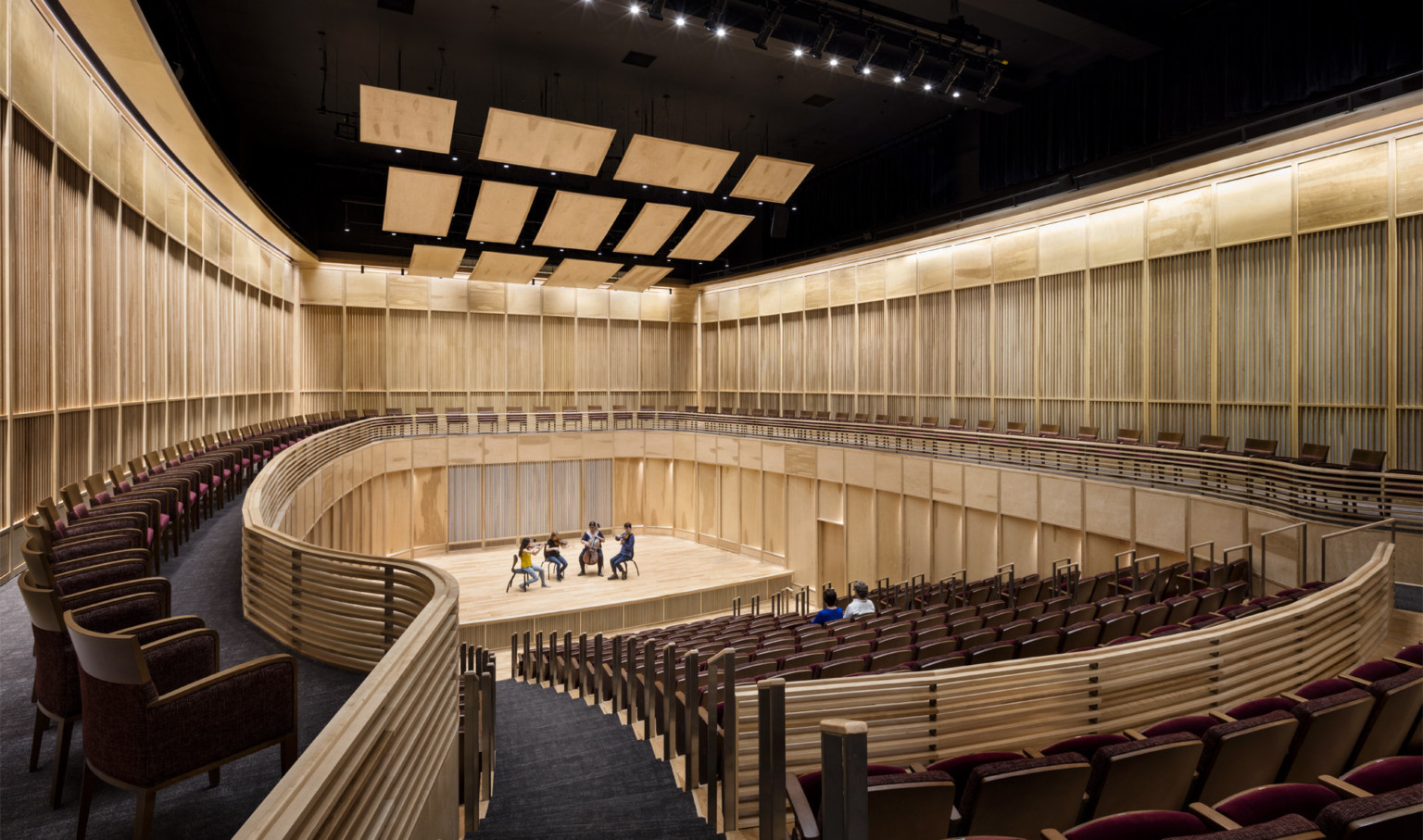 View from the back of wooden wall oval auditorium with 4 musicians on stage and 2 people seated in the audience View from the back of wooden wall oval auditorium with 4 musicians on stage and 2 people seated in the audience