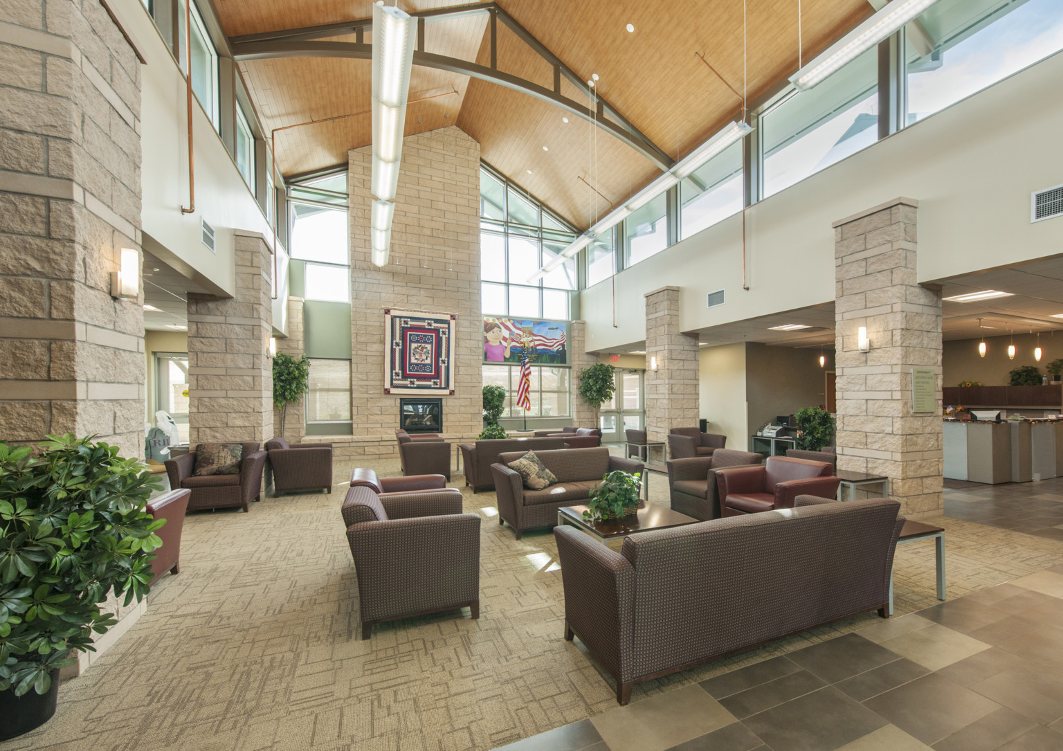 A view of the main room in the soldier family assistance center, brown tile and carpet with burgundy sofas and chairs, tan stone columns support neutral walls with clerestory windows provide daylight views, plants promote harmony