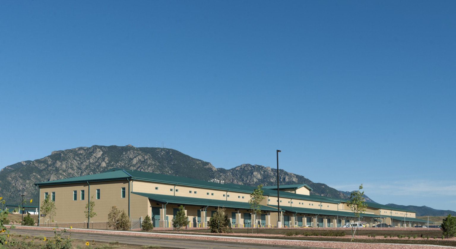 Two story yellow CMU building with green metal roof and loading docks surrounded by secure fence. Mountains in background