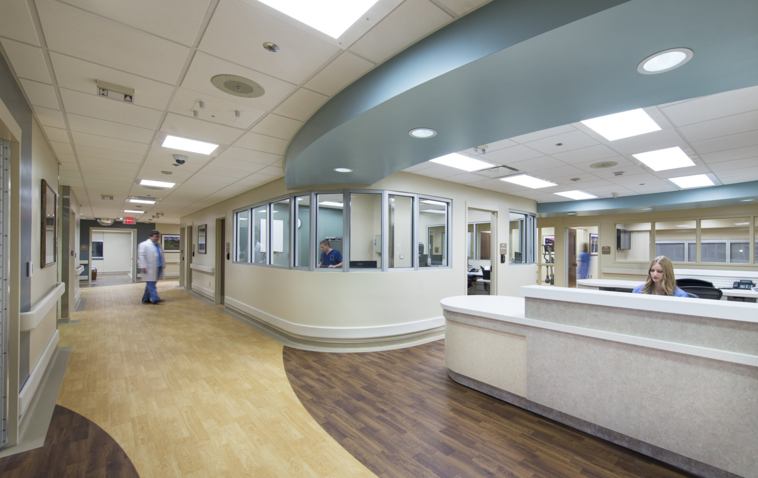 Hospital with wood flooring and neutral walls. Blue ceiling soffit curves over public walkway lined with glass front offices.