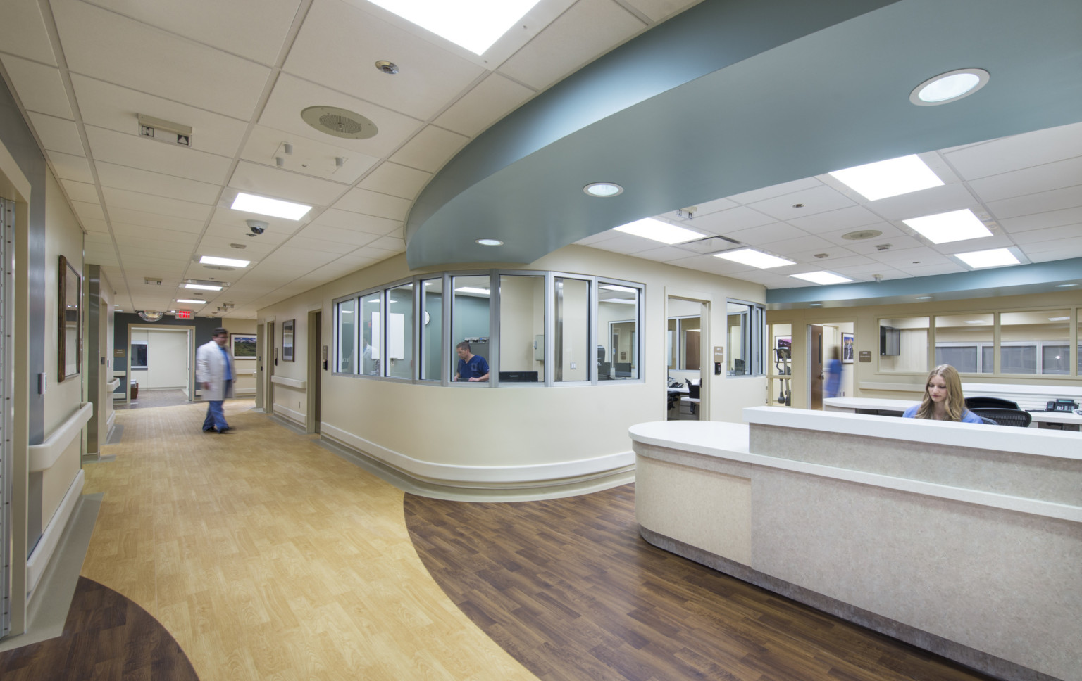 Hospital with wood flooring and neutral walls. Blue ceiling soffit curves over public walkway lined with glass front offices. Hospital with wood flooring and neutral walls. Blue ceiling soffit curves over public walkway lined with glass front offices.