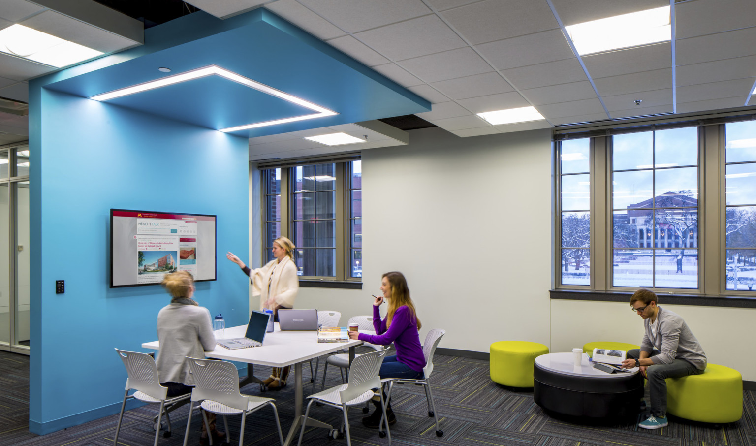 small group student work at a presentation screen under blue wrapped ceiling accent which goes to floor. Illuminated above small group student work at a presentation screen under blue wrapped ceiling accent which goes to floor. Illuminated above
