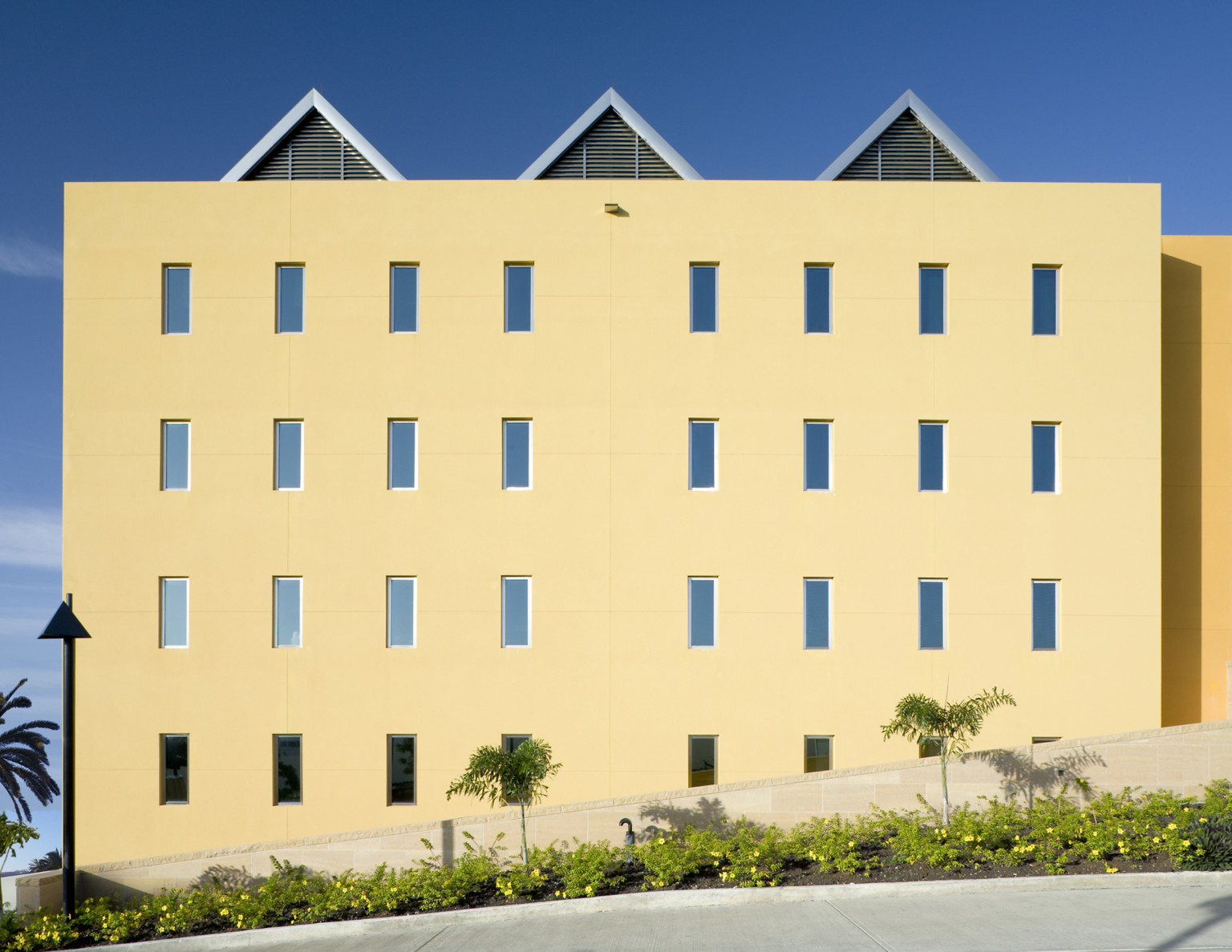 square yellow building with evenly repeating rectangular windows and palm trees. Three peaked roofs spaced evenly on top square yellow building with evenly repeating rectangular windows and palm trees. Three peaked roofs spaced evenly on top