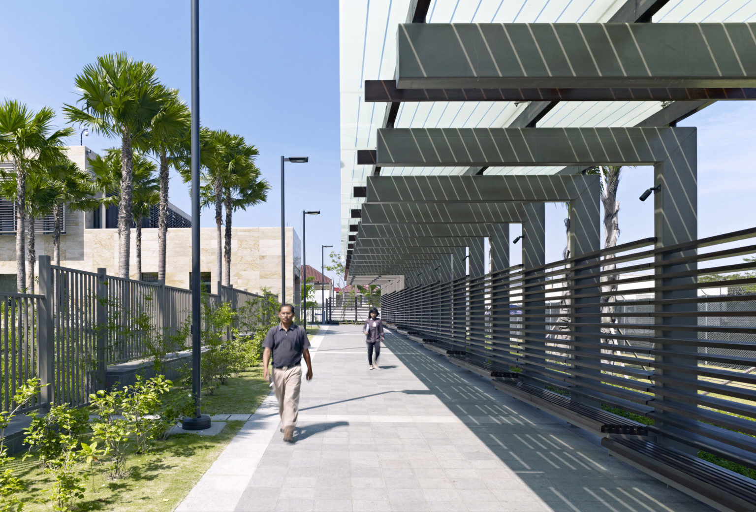 concrete brick path lined with vertical and horizontal fencing. Perimeter has transparent canopy and slatted benches.