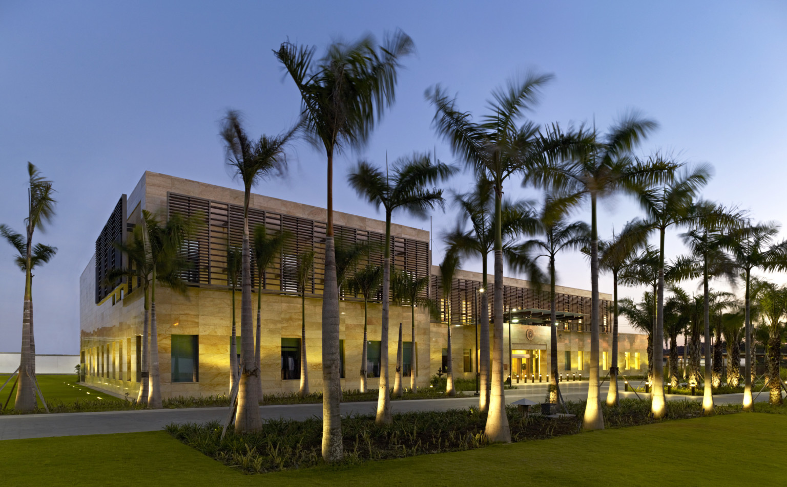 a stone building with brise soleil panels on second story windows. uplights illuminate ground floor, windows, and palm trees
