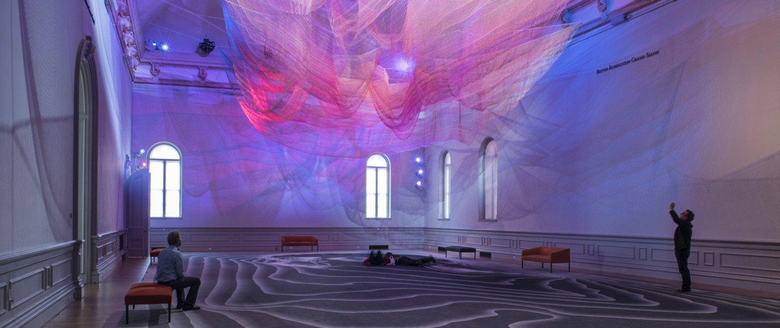 Colorful abstract art installation with pink and purple light on layers of sheer netting with shadows and light on white wall